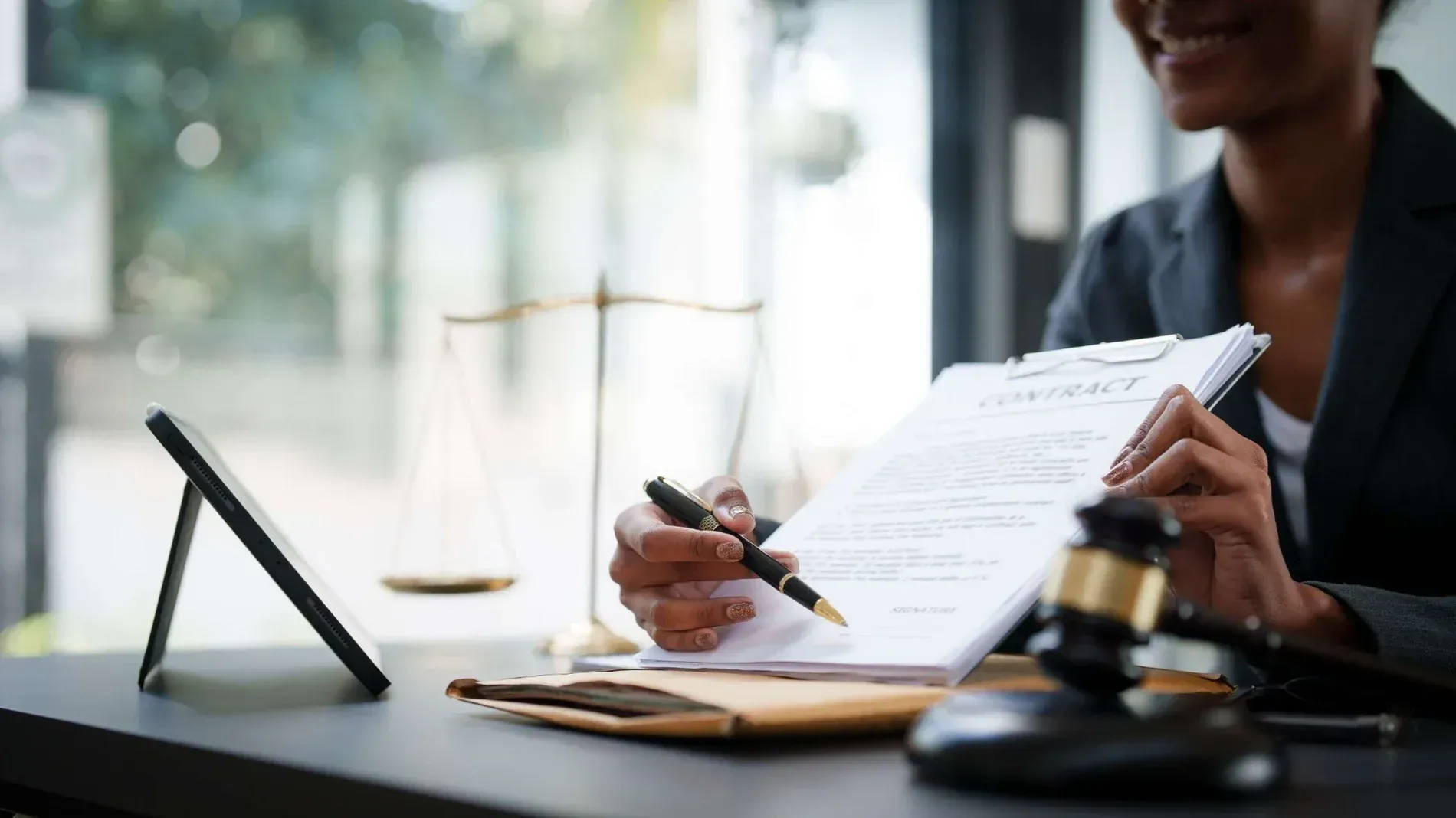 Un abogado señala un contrato en un portapapeles. La balanza de la justicia, el mazo y la tablilla están sobre el escritorio.