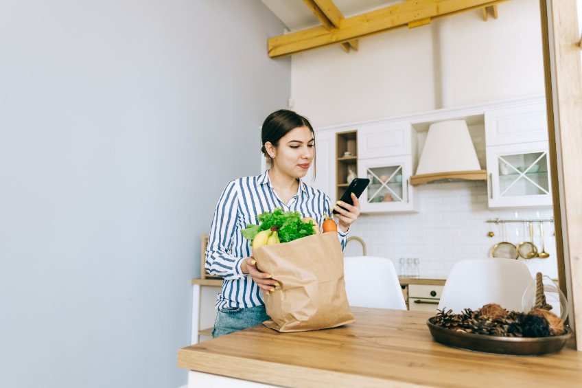 Una mujer sostiene una bolsa de comestibles y mira su teléfono.
