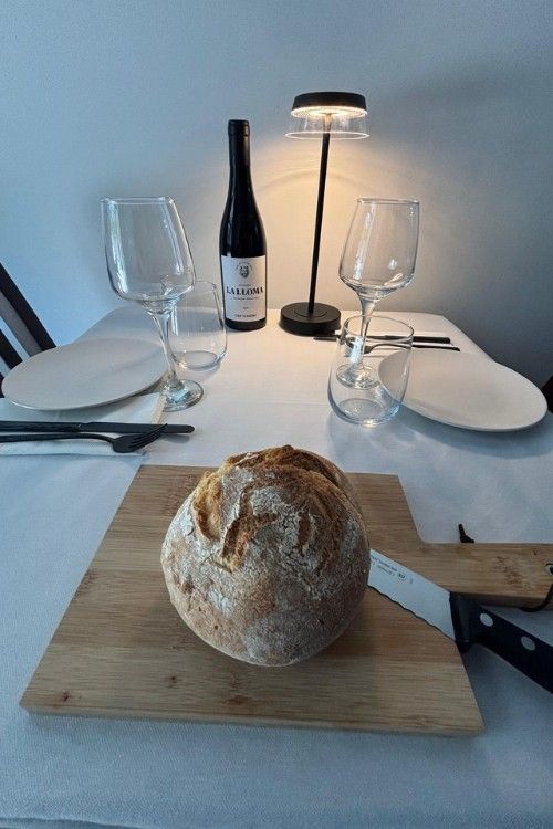 Mesa puesta para la cena, incluyendo pan sobre tabla de madera, vino, copas, platos y lámpara.