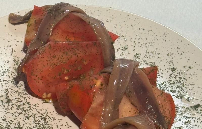 Rodajas de tomate cubiertas con anchoas, condimentadas con hierbas, en un plato blanco.
