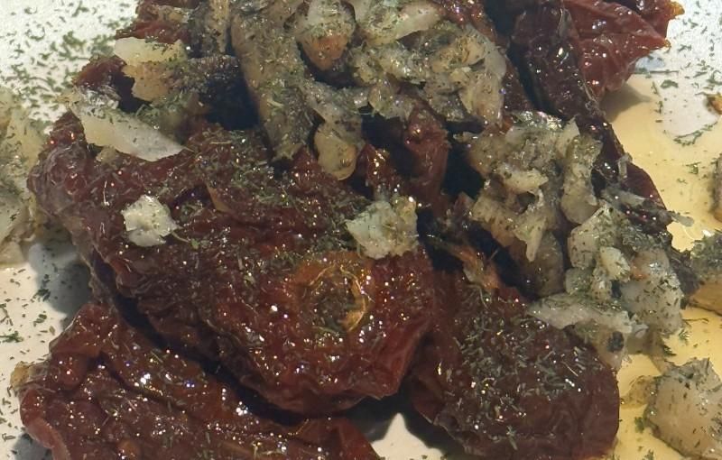 Tomates secados al sol cubiertos con cebollas picadas y hierbas, en un plato blanco.