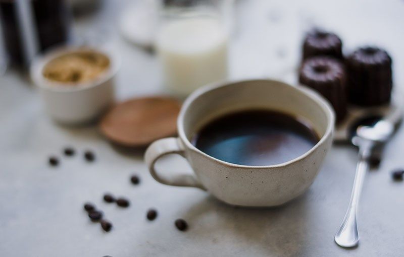 Café en una taza moteada, cerca de granos de café, pequeños pasteles, azúcar, leche y una cuchara sobre una superficie clara.
