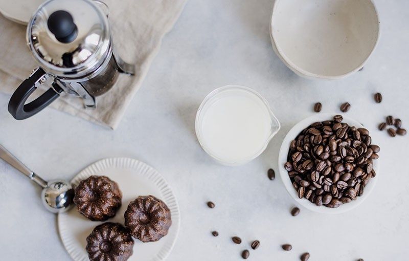Prensa francesa, pasteles, granos de café y leche sobre una superficie blanca.