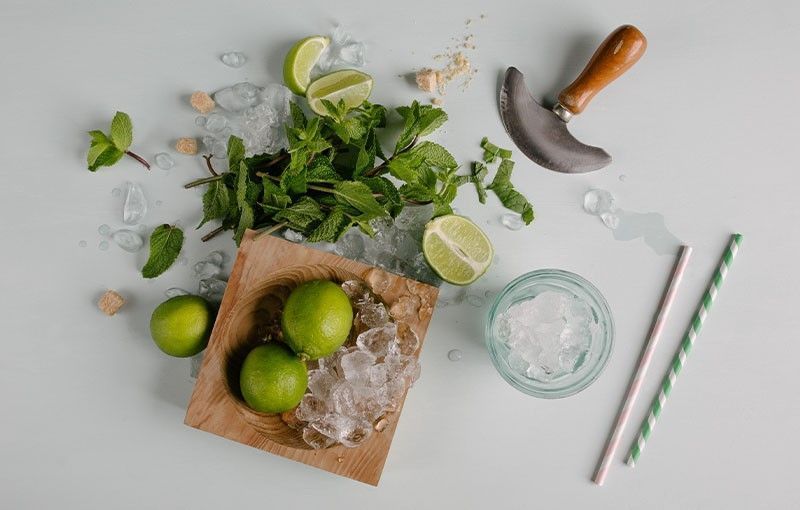 Ingredientes del mojito: limas, menta, hielo, una mezzaluna y pajitas sobre una superficie azul claro.