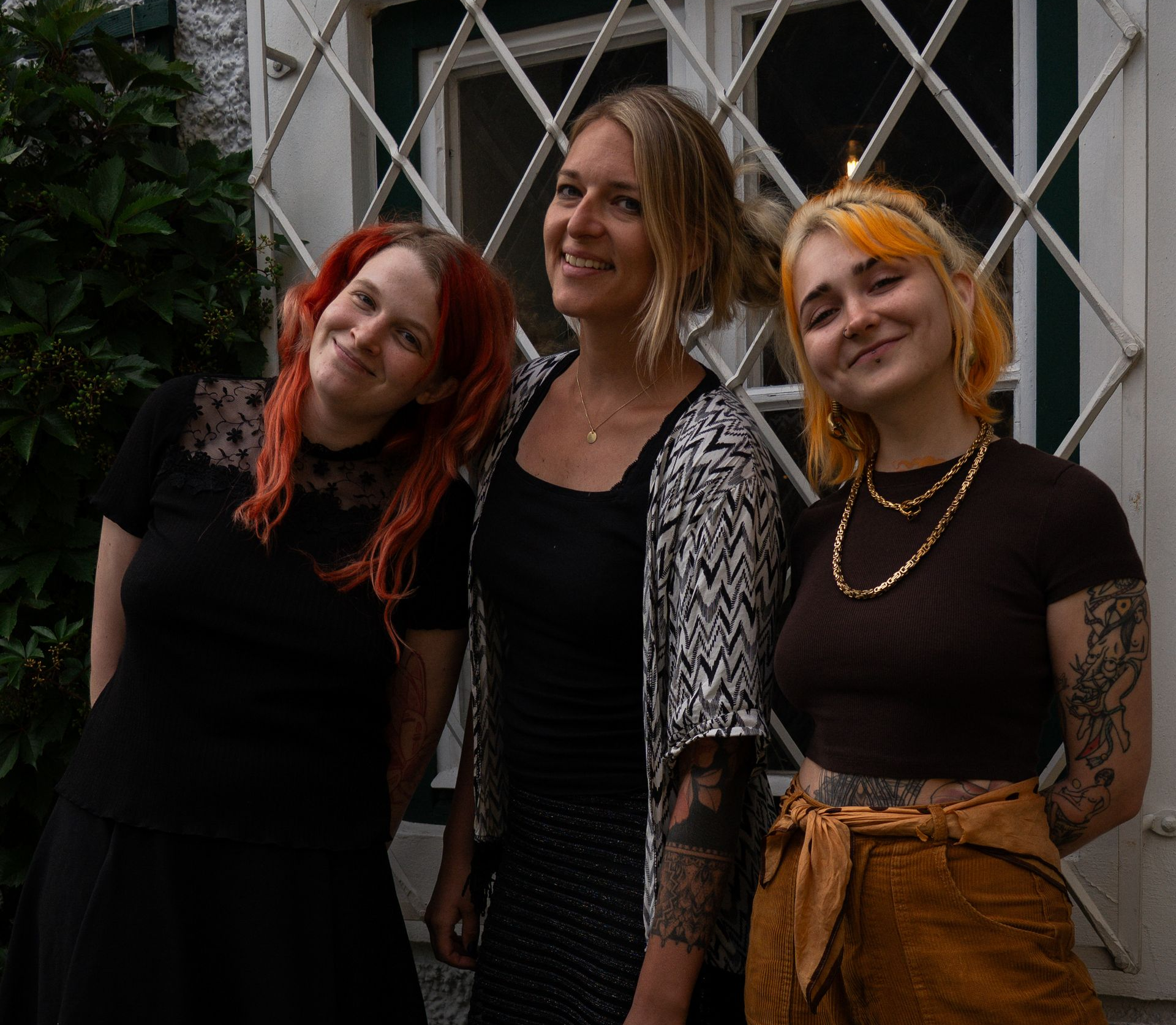 Drei lächelnde Frauen, umgeben von gerahmter Kunst. Eine Frau hat Tätowierungen auf Armen und Brust.