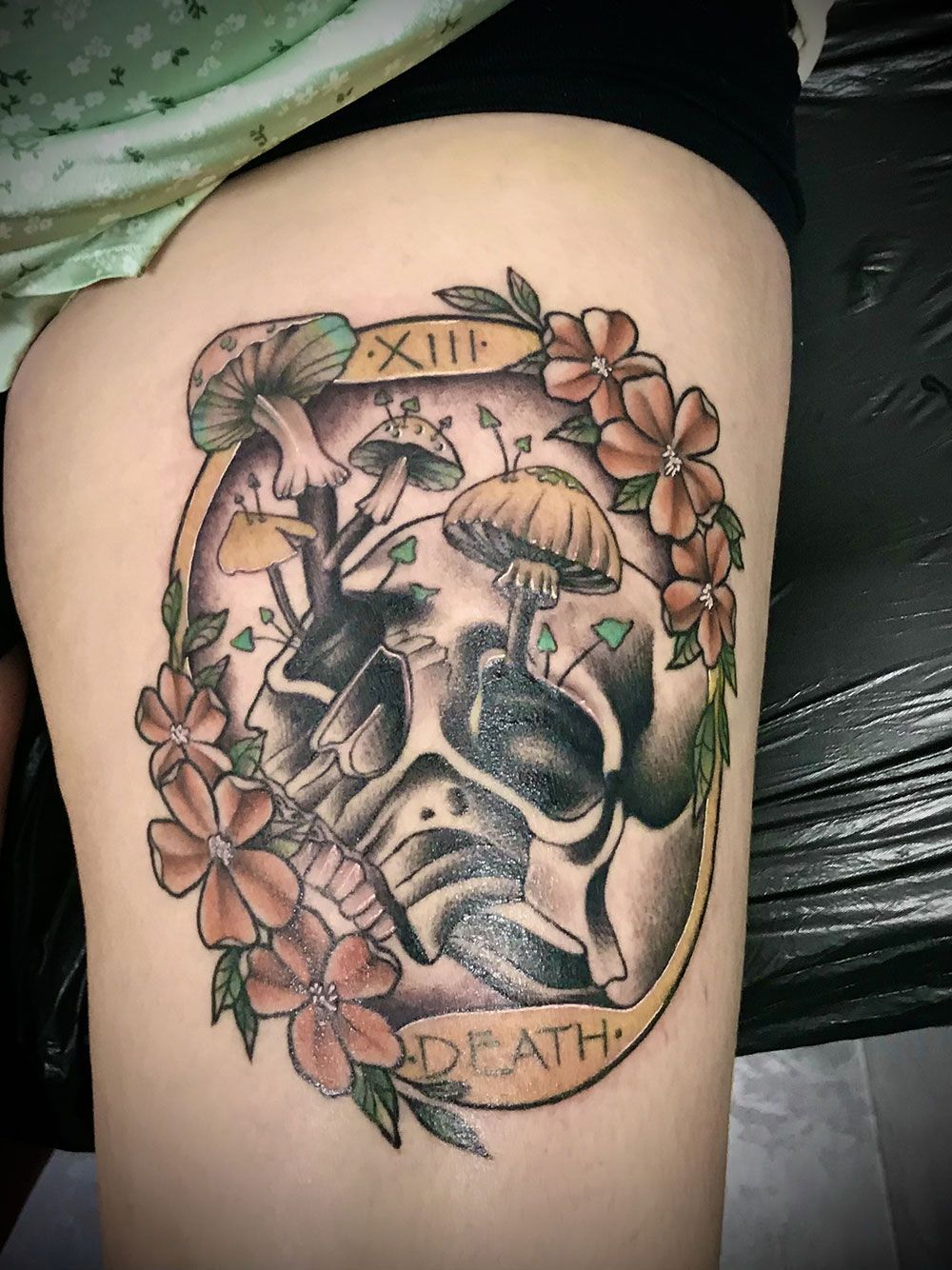 Eine Frau hat ein Tattoo eines Totenkopfes mit Pilzen und Blumen auf ihrem Bein