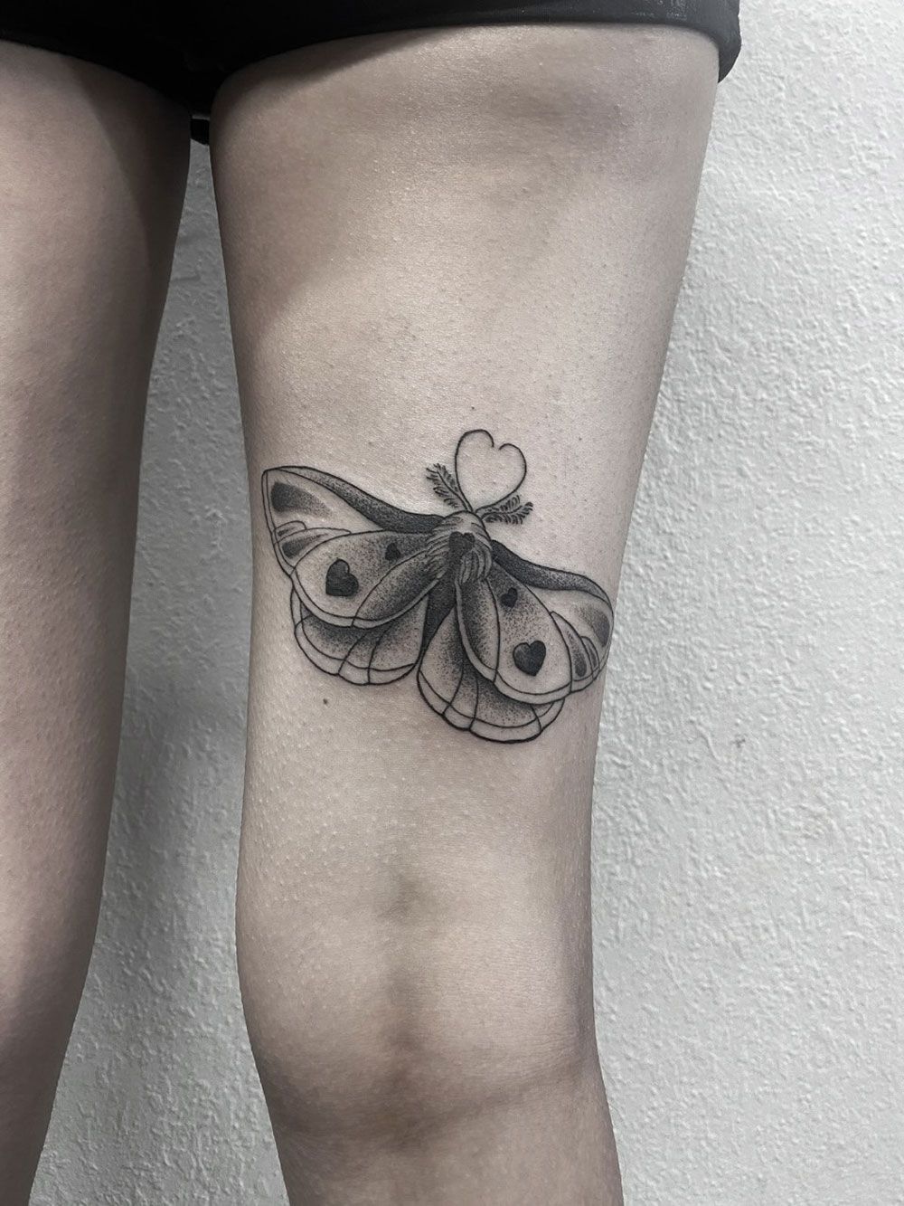 Eine schwarz-weiße Tätowierung einer Motte mit einem Herz auf den Flügeln auf dem Bein einer Frau.