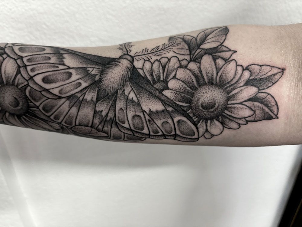 Eine schwarz-weiße Tätowierung eines Schmetterlings und von Blumen auf dem Arm einer Person.