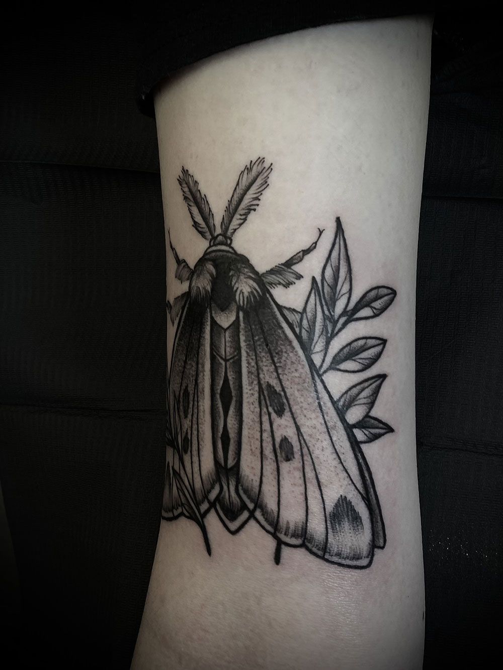 Eine schwarz-weiße Tätowierung einer Motte mit Blättern auf dem Bein einer Person.