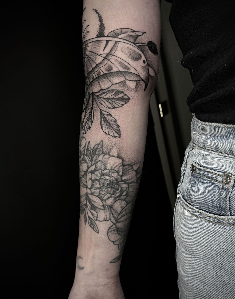 Eine Frau hat eine Tätowierung mit einer Schildkröte und Blumen auf ihrem Unterarm.