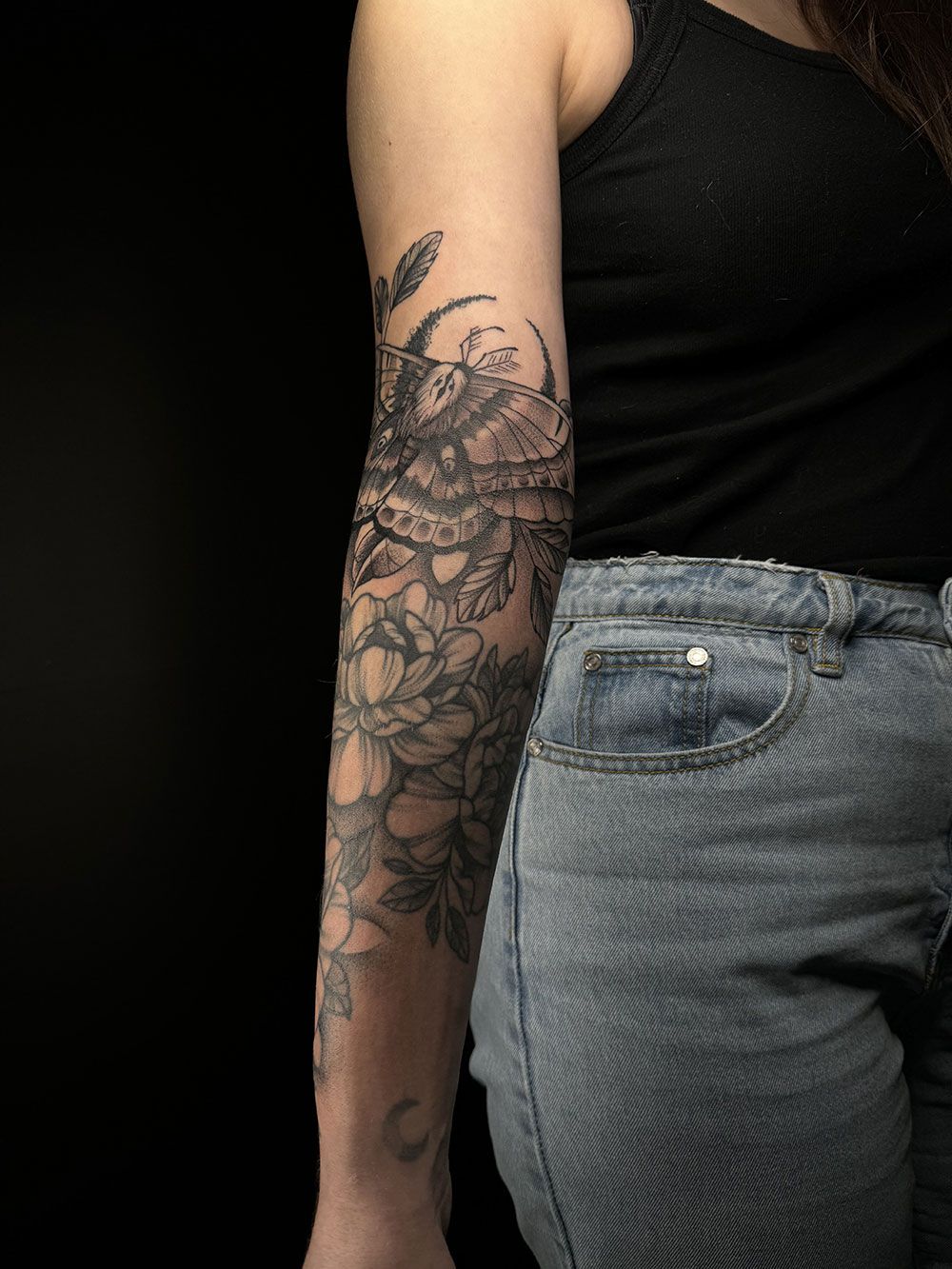 Eine Frau mit einer Tätowierung auf dem Arm trägt Jeans und ein schwarzes Tanktop.