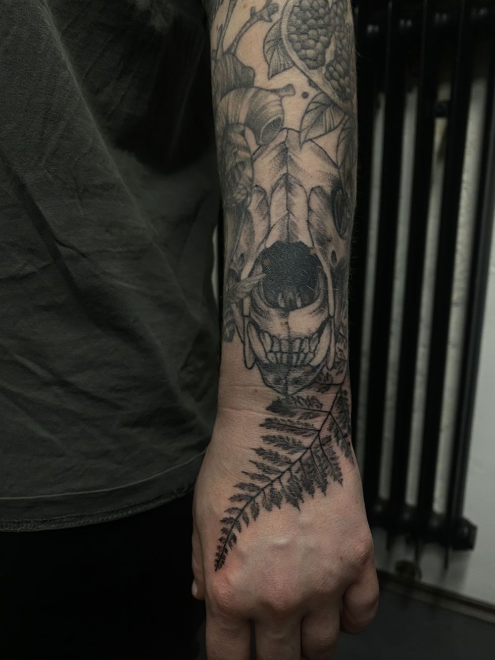 Eine Person hat eine Tätowierung mit einem Totenkopf und einem Farn auf dem Unterarm.