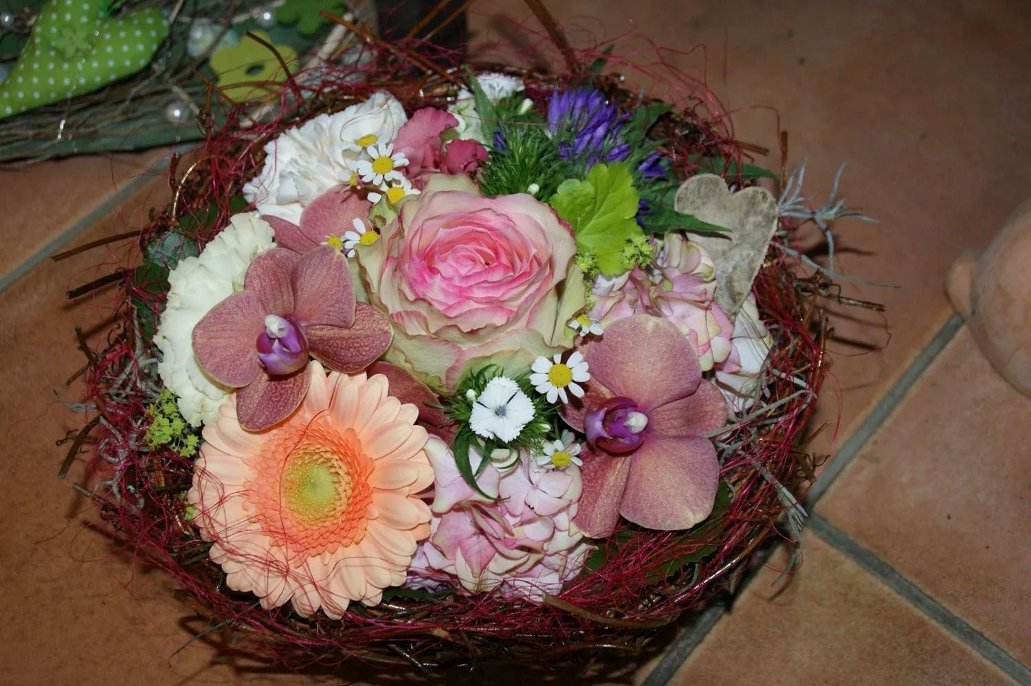 Blumenarrangement mit rosa, weissen und orangefarbenen Blüten, umgeben von einer rot gewebten Bordüre, auf einer gefliesten Oberfläche.