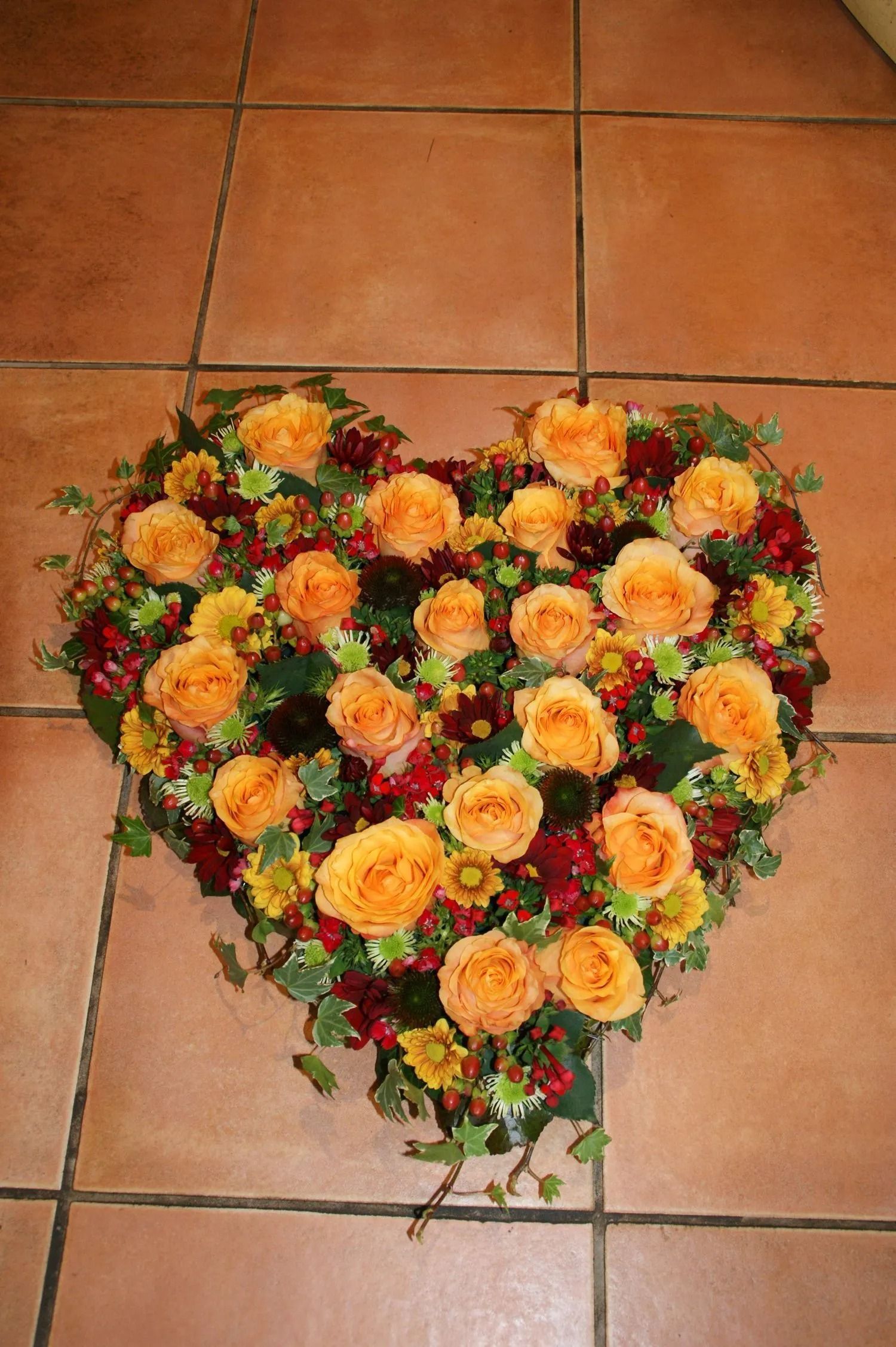 Herzförmiges Blumenarrangement; orange Rosen, rote und gelbe Akzente; brauner Fliesenboden.