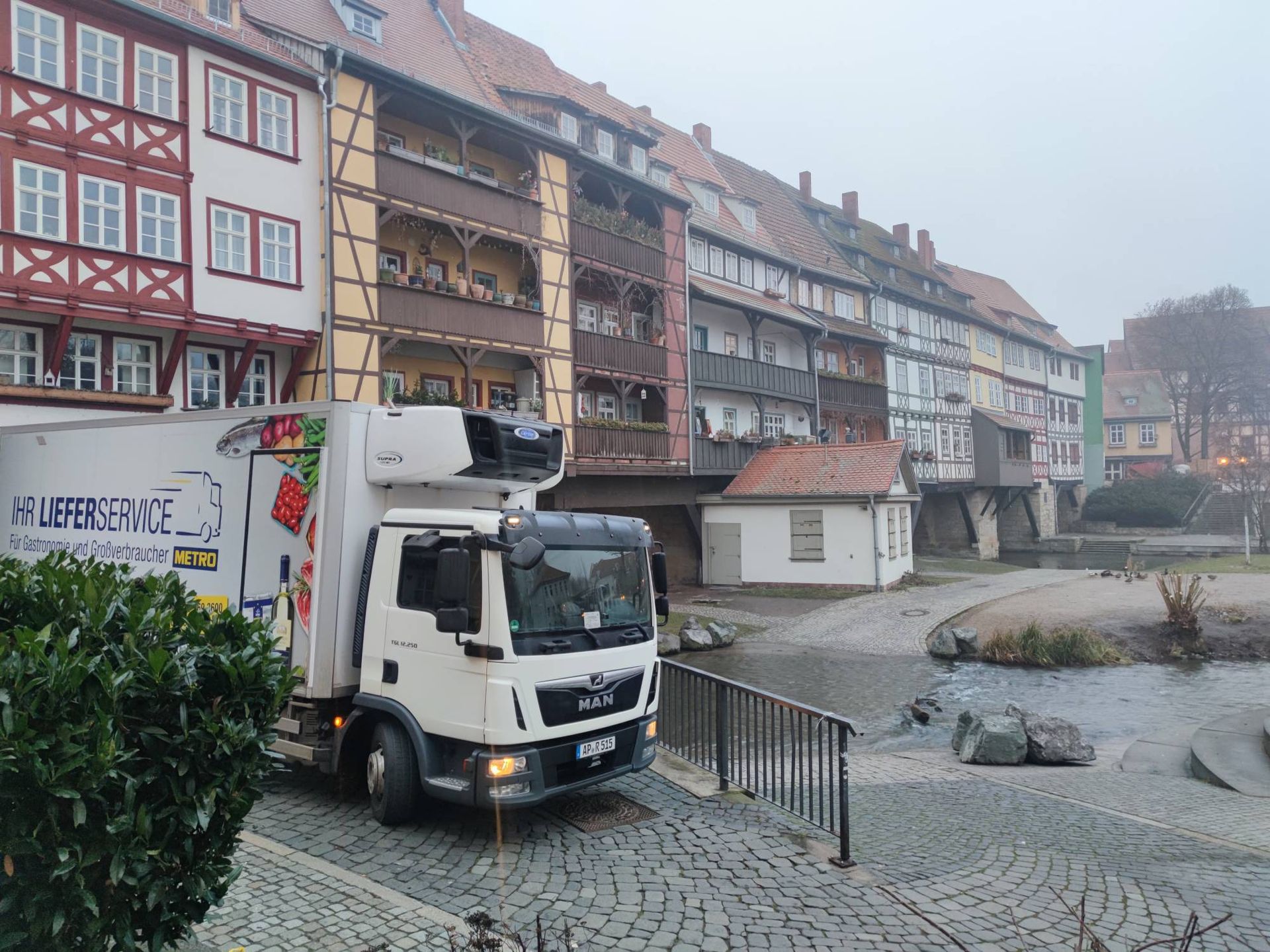 Kühlwagen auf Kopfsteinpflasterstraße, neben Bach und Fachwerkhäusern. Bewölkter Tag.