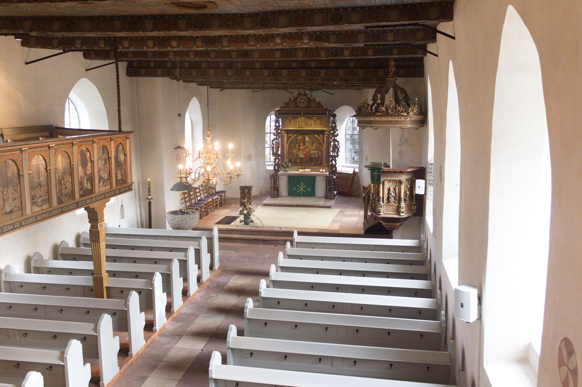 Der Innenraum einer Kirche. Zu sehen sind u.a. hellgraue Bankreihen, eine geschnitzte Kanzel und ein Altar mit Retabel.