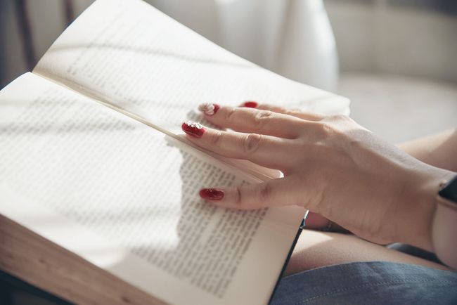 Mujer con uñas pintadas de rojo sosteniendo un libro abierto; la luz del sol ilumina las páginas.