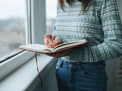 Una persona escribe en un cuaderno cerca de una ventana, viste un suéter azul estampado y jeans.