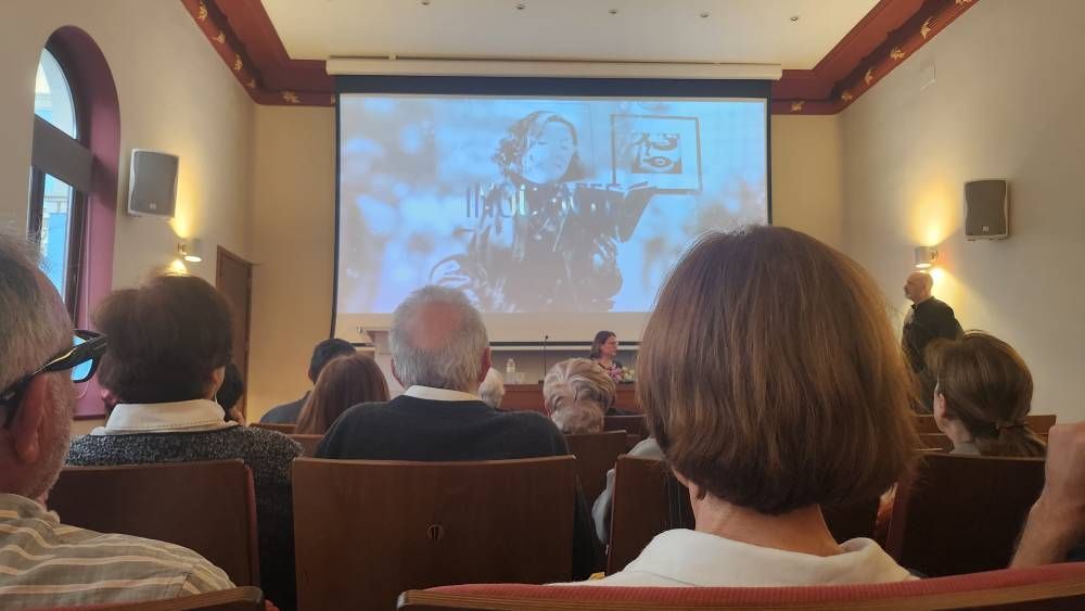 El público observa la presentación en pantalla en una sala. El orador está en el podio. La imagen muestra a una persona sosteniendo una foto.