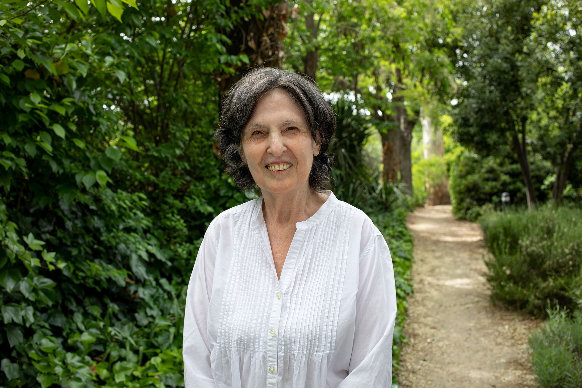 Una mujer de cabello oscuro y camisa blanca sonríe en un jardín con follaje verde y un camino.