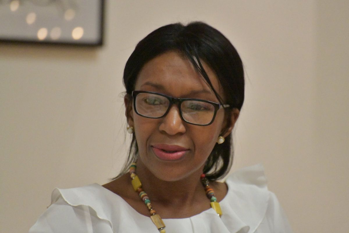 Mujer con gafas, vestida con una blusa blanca y un collar de cuentas de colores en una habitación con un fondo neutro.
