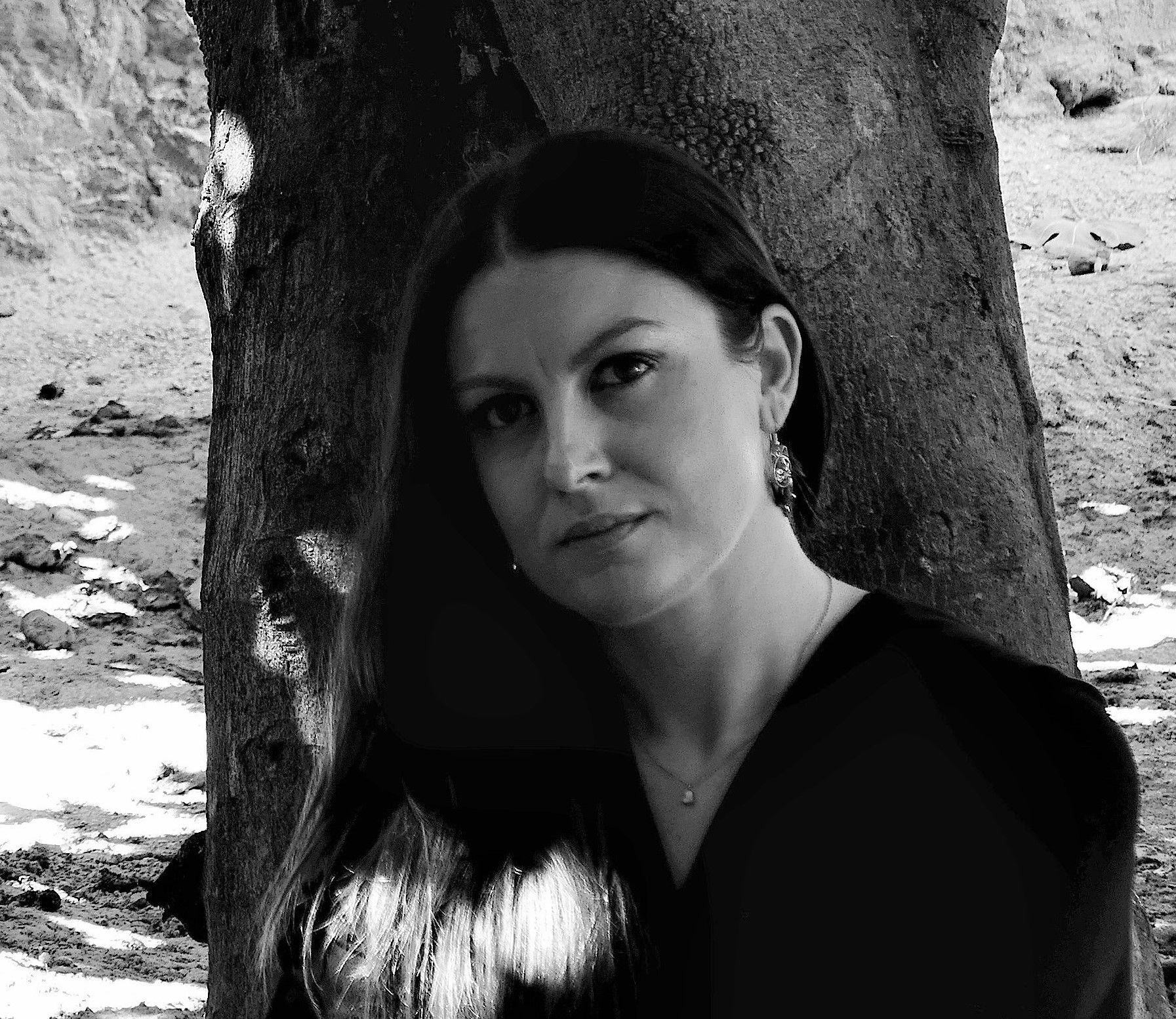 Mujer con cabello largo y oscuro, vestida con ropa oscura, se apoya contra el tronco de un árbol en un entorno natural.
