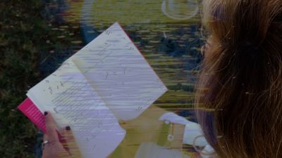 Una persona escribe en un cuaderno cerca de una ventana, viste un suéter azul estampado y jeans.