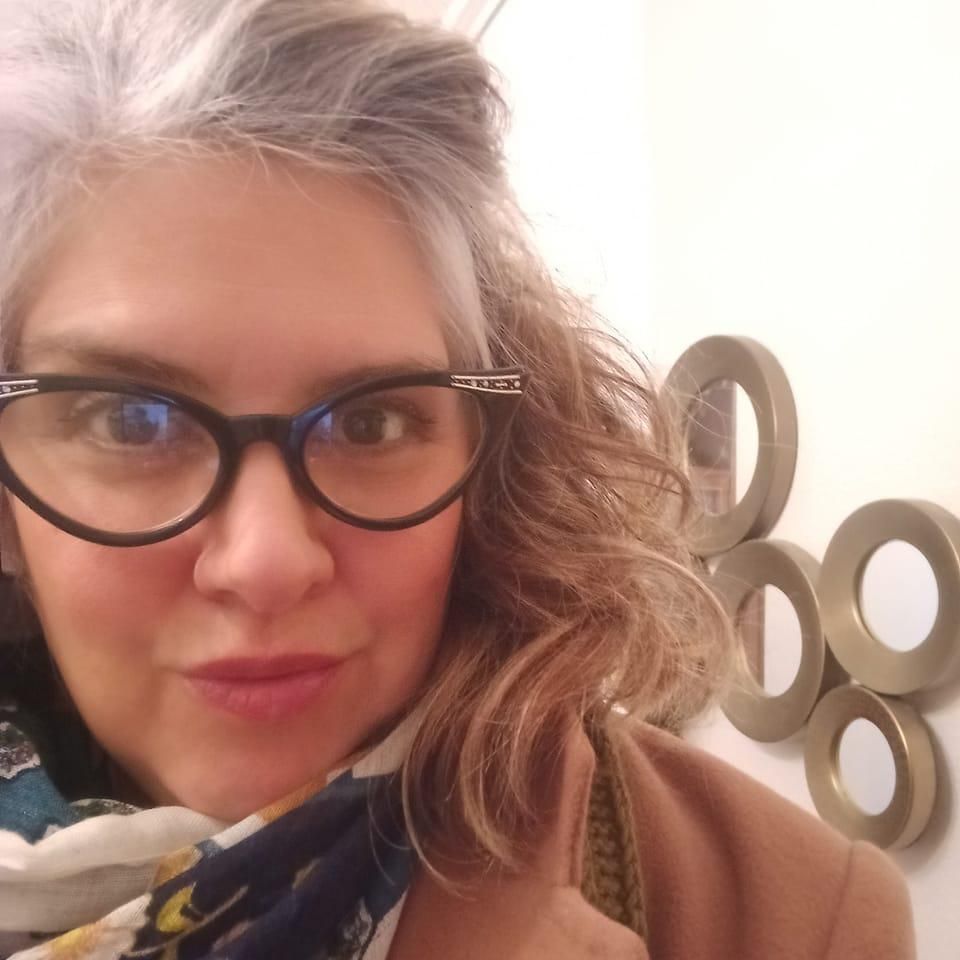 Mujer con cabello canoso y gafas sonríe, cerca de una decoración de pared dorada.