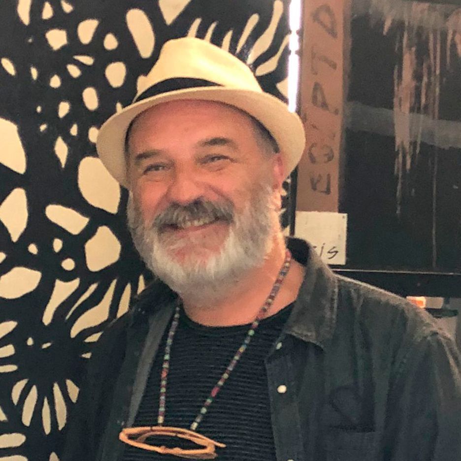Un hombre con barba gris y sombrero sonríe frente a una pared con dibujos en blanco y negro.