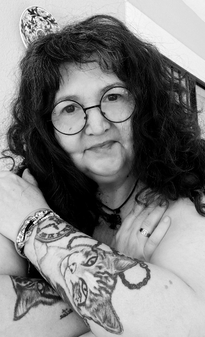 Mujer con cabello oscuro y rizado, gafas y tatuajes en los brazos, sonriendo, con los brazos cruzados.
