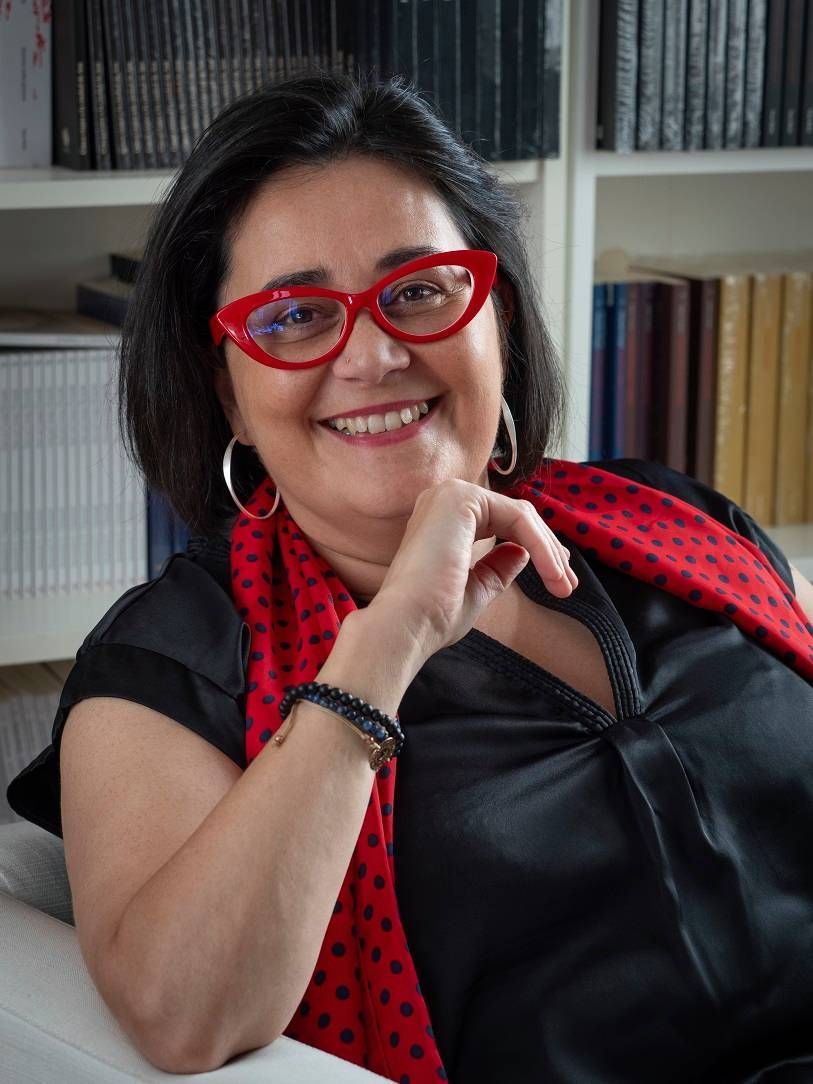 Mujer con gafas rojas sonríe, apoya la barbilla en la mano; estanterías de libros en el fondo.