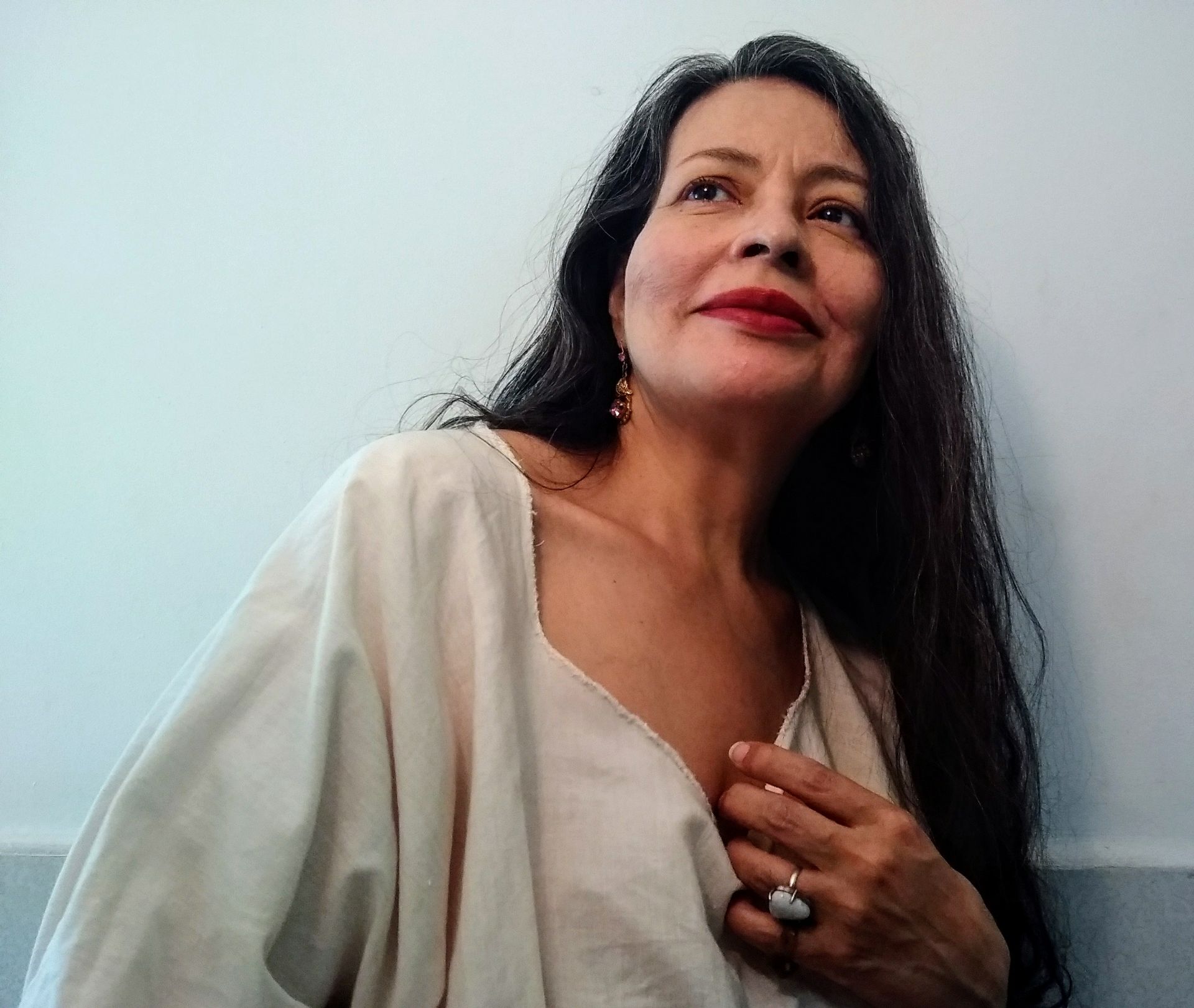 Una mujer de cabello oscuro y camisa blanca sonríe en un jardín con follaje verde y un camino.