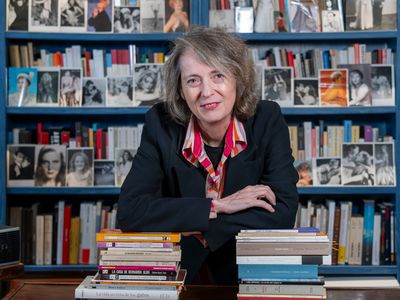 Mujer sentada en un escritorio con pilas de libros, sonriendo frente a una estantería.