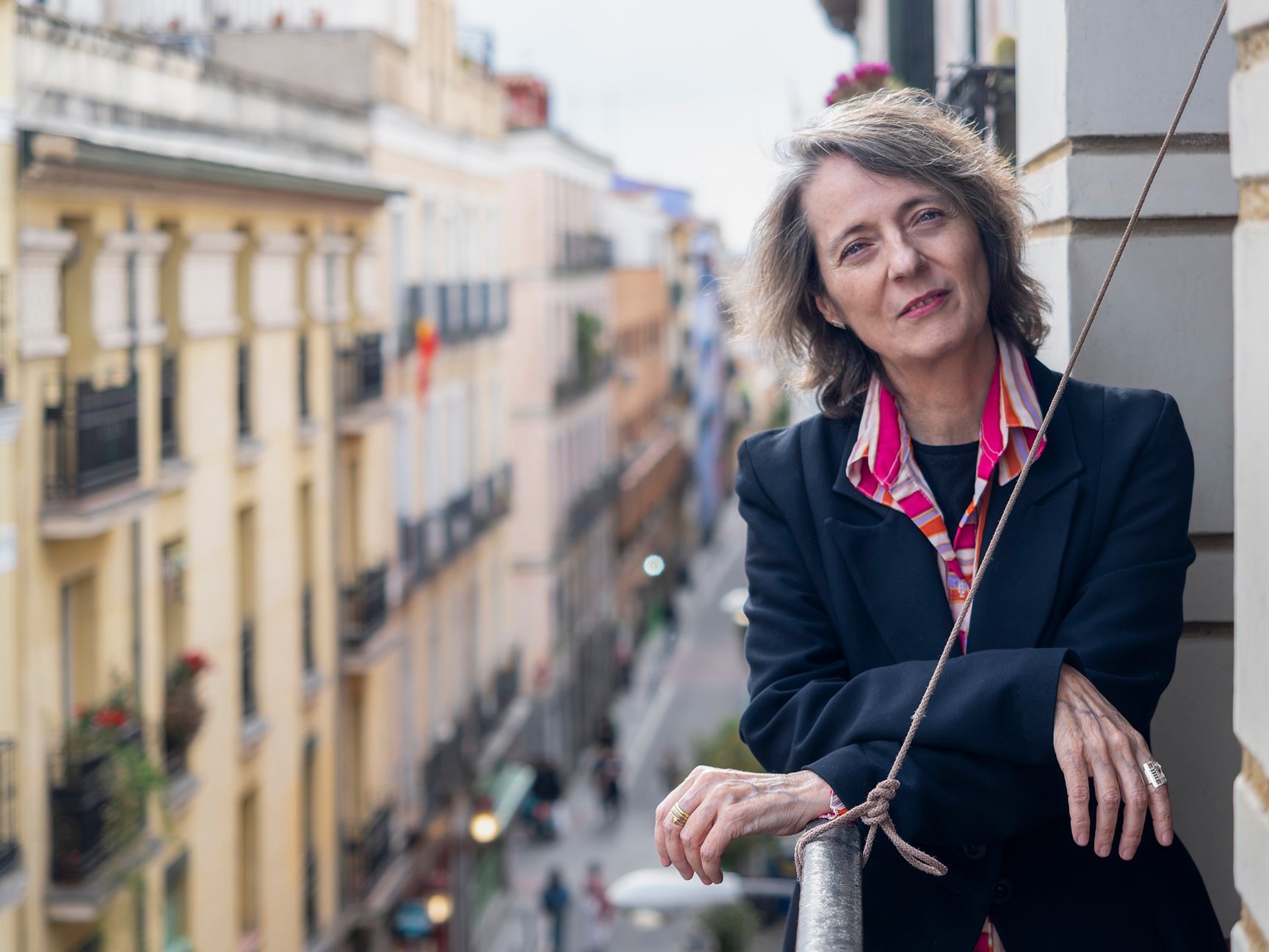 Mujer con blazer se apoya en un balcón, con vista a una calle de la ciudad.