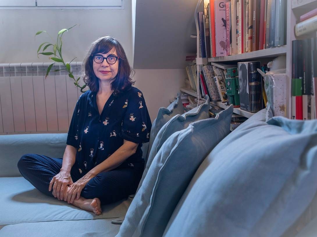 Mujer sentada con las piernas cruzadas en un sofá en un interior, con gafas, mirando a la cámara. Estantería visible.