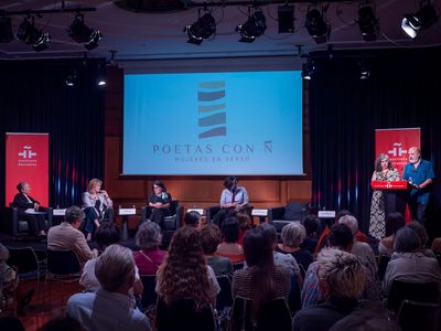 Público en una lectura de poesía; escenario iluminado con una pantalla que muestra 