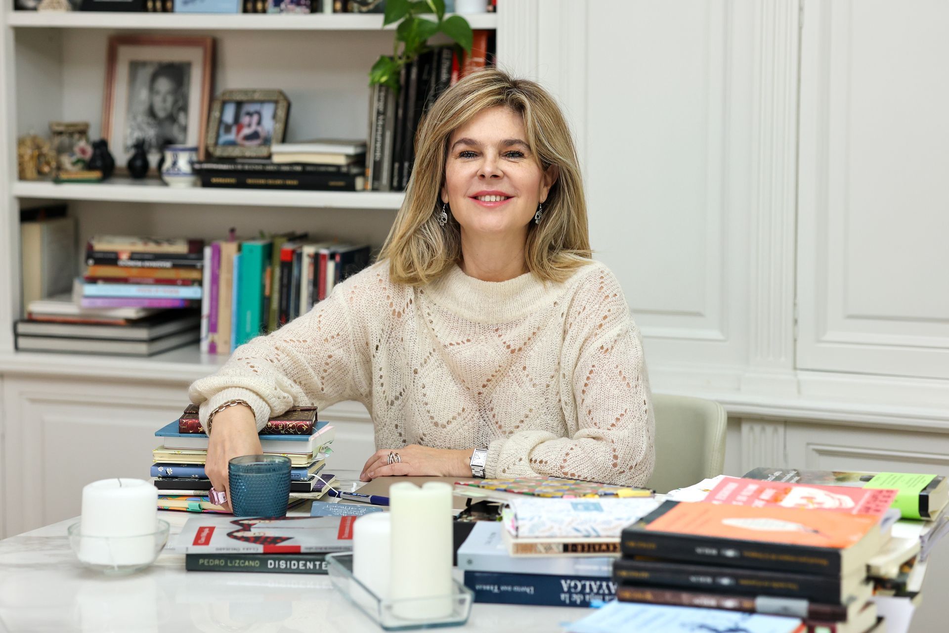 Mujer en una mesa con libros, sonriendo, vistiendo un suéter color crema, estanterías de fondo.