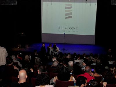 Público en una lectura de poesía; escenario iluminado con una pantalla que muestra
