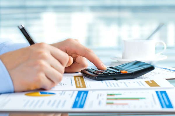 Manos usando una calculadora sobre documentos financieros con gráficos, junto a una taza de café sobre un escritorio.