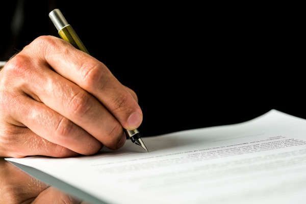 Una mano sostiene una pluma estilográfica, lista para firmar un documento sobre un escritorio reflectante con fondo negro.