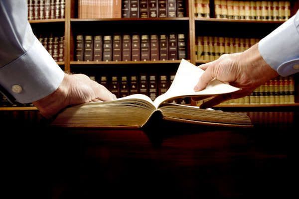 Dos personas vestidas con camisas de vestir abren un libro grande y grueso sobre un escritorio de madera frente a una estantería de la biblioteca llena de libros.