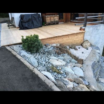 Une terrasse en bois est entourée de rochers et de plantes devant une maison.