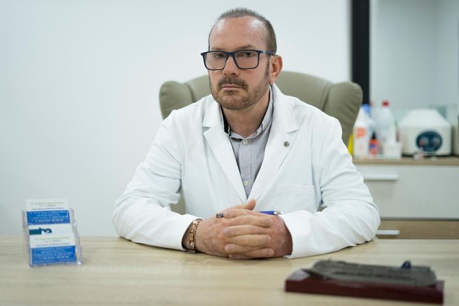 Un hombre con una bata de laboratorio está sentado en un escritorio con las manos cruzadas.