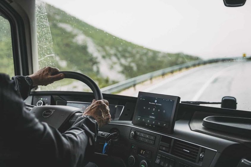 Las manos del conductor sobre el volante de un camión, conduciendo por una carretera mojada, con montañas al fondo.