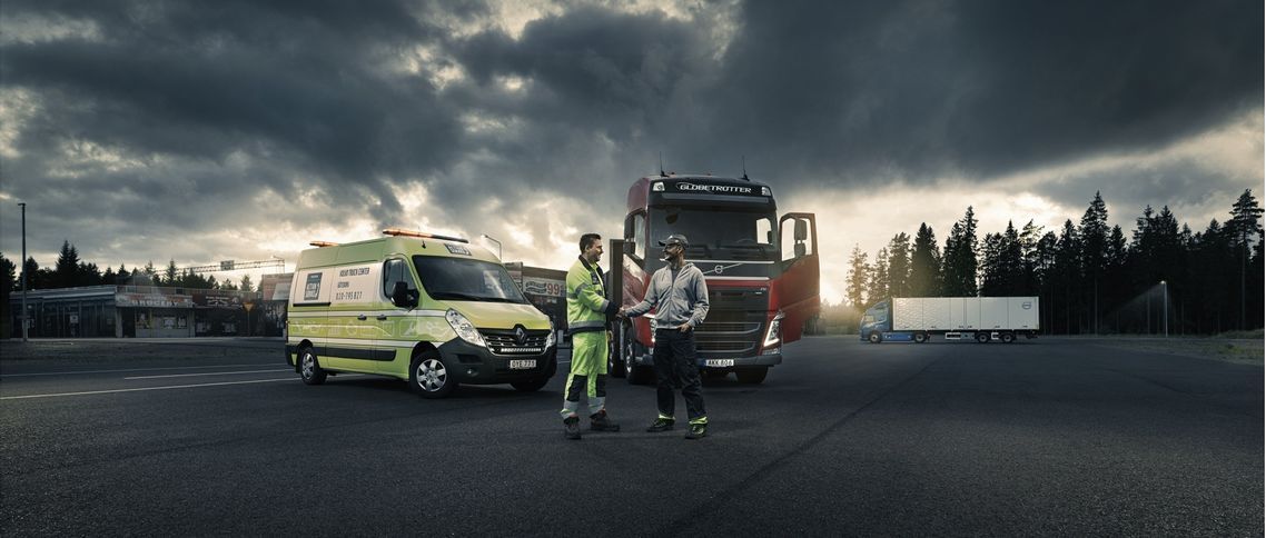 Ambulancia y camión semirremolque con dos personas de pie sobre un terreno asfaltado oscuro bajo un cielo nublado.