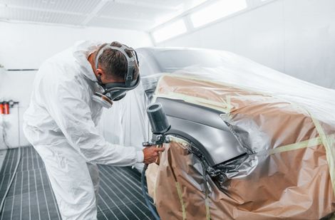 Ein Arbeiter lackiert eine Autokarosserie in einer mit Schutzfolie abgedeckten Autowerkstatt.