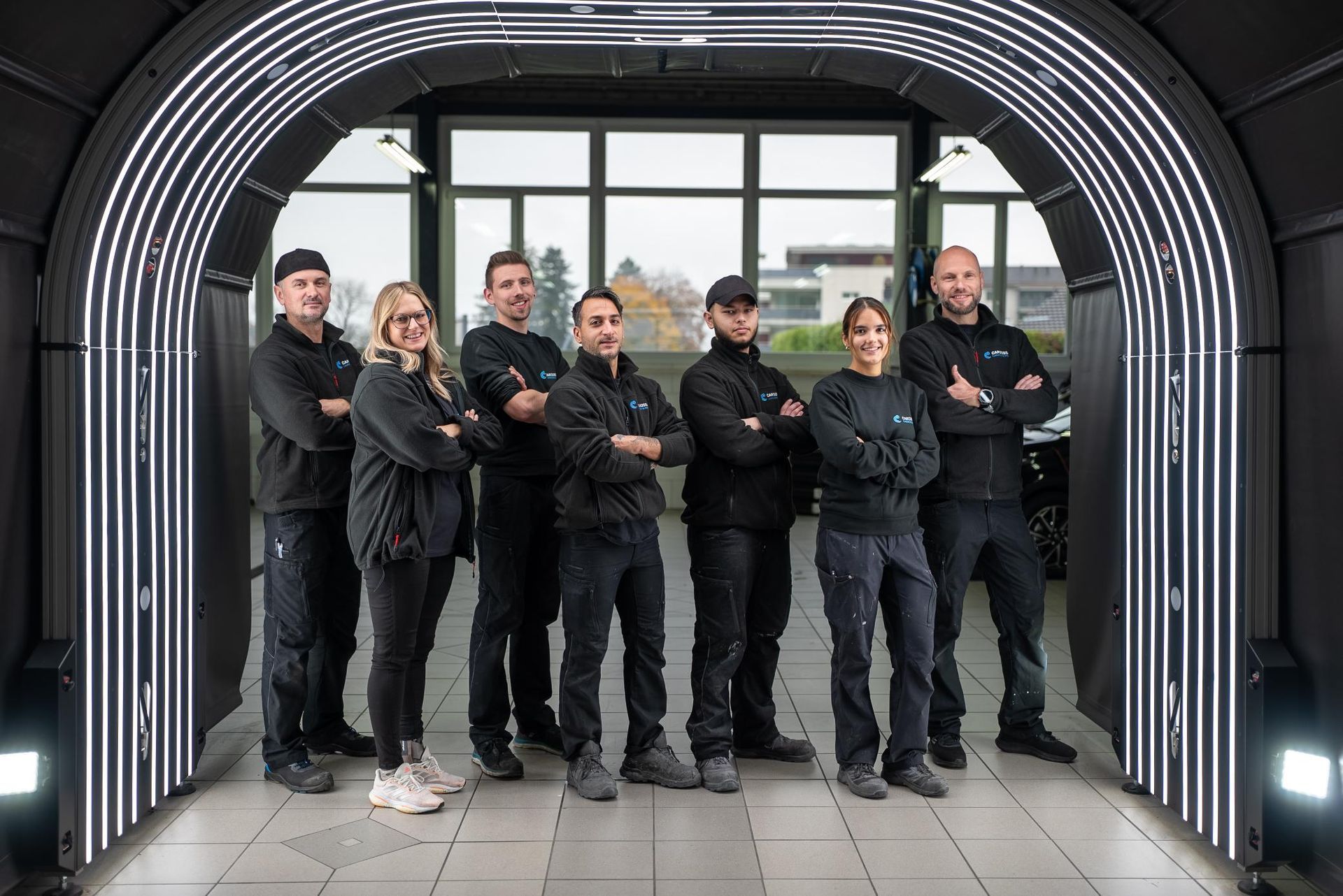 Sechs Personen in schwarzer Kleidung stehen mit verschränkten Armen in einem modernen Torbogen.