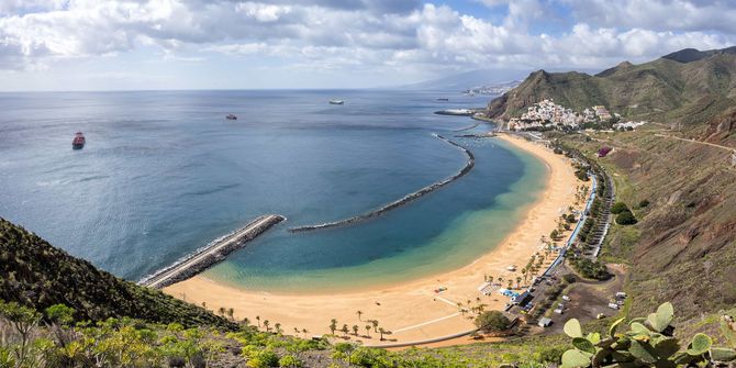 Vue aérienne d'une plage en forme de croissant aux eaux turquoise, de collines rocheuses et d'une digue s'avançant dans la baie