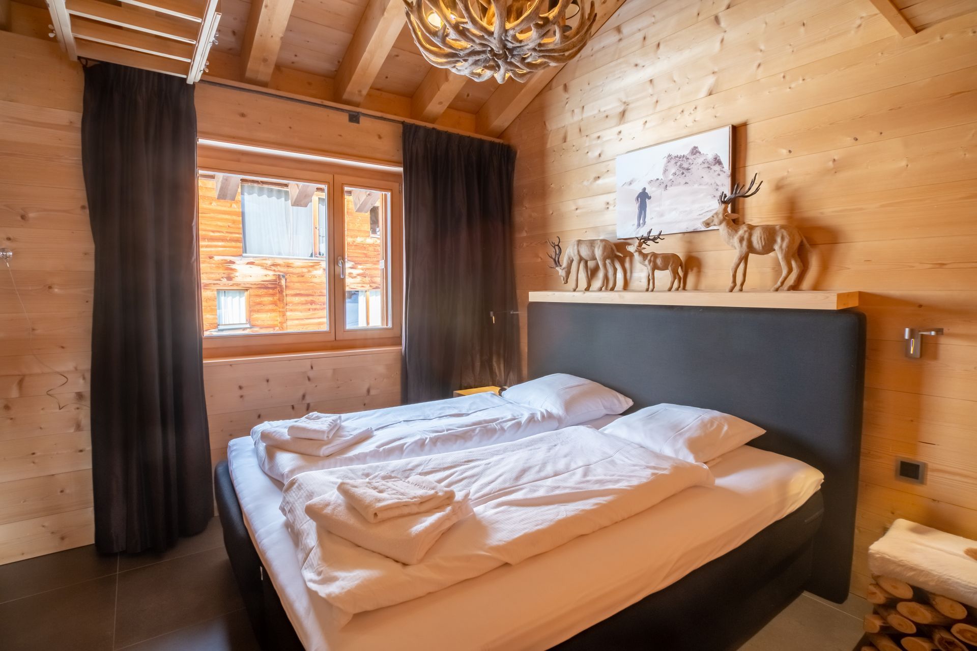 Schlafzimmer mit Holzwänden, Bett mit weißer Bettwäsche, Fenster mit dunklen Vorhängen und dekorativem Rentier.