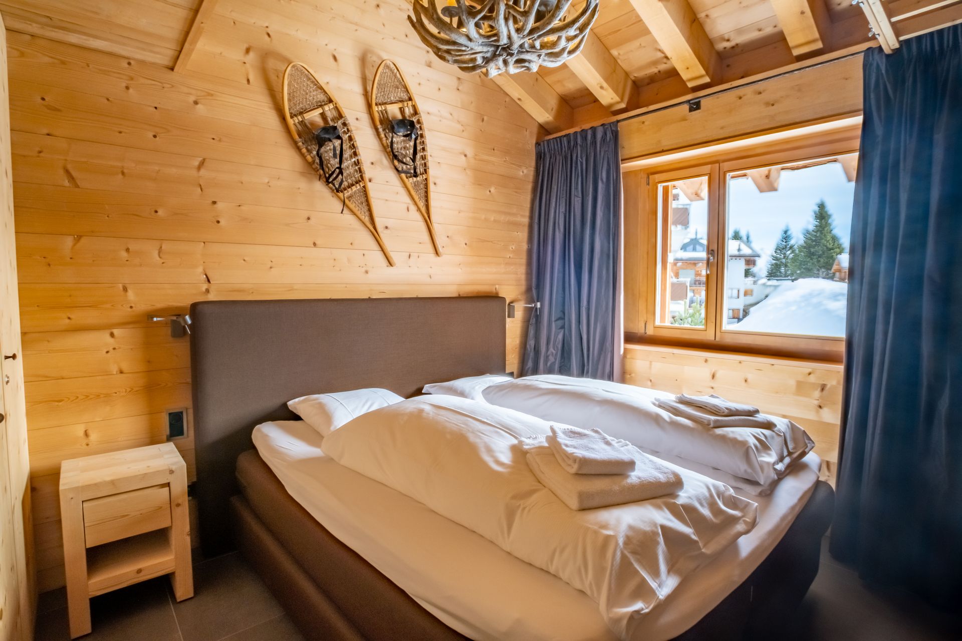 Schlafzimmer mit Holzwänden, Bett, Nachttisch, Schneeschuhen und einem Fenster mit Blick auf die verschneite Landschaft.